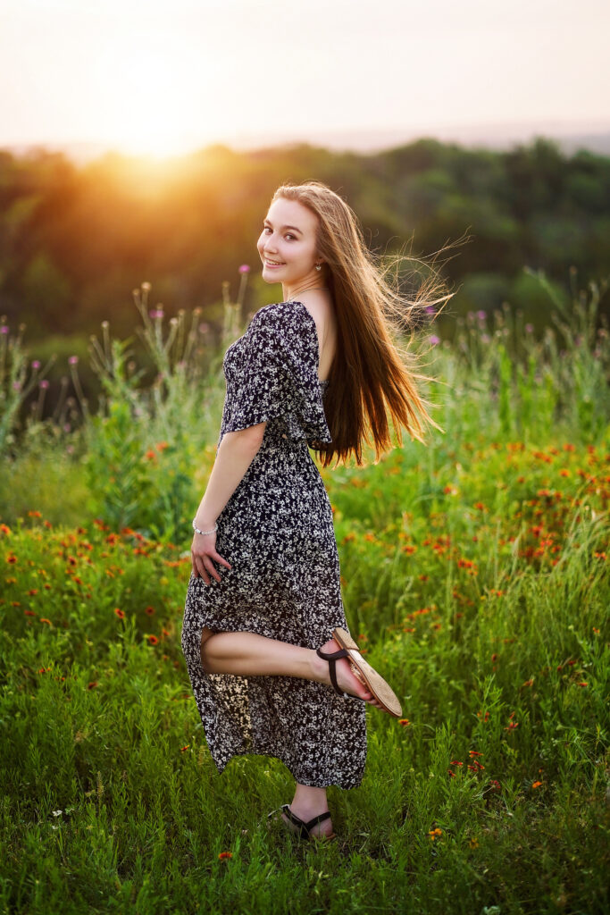 Jaelyn smiling in a sunlit field of wildflowers, wearing a flowy dress with one foot lifted playfully as the sun sets behind her.
