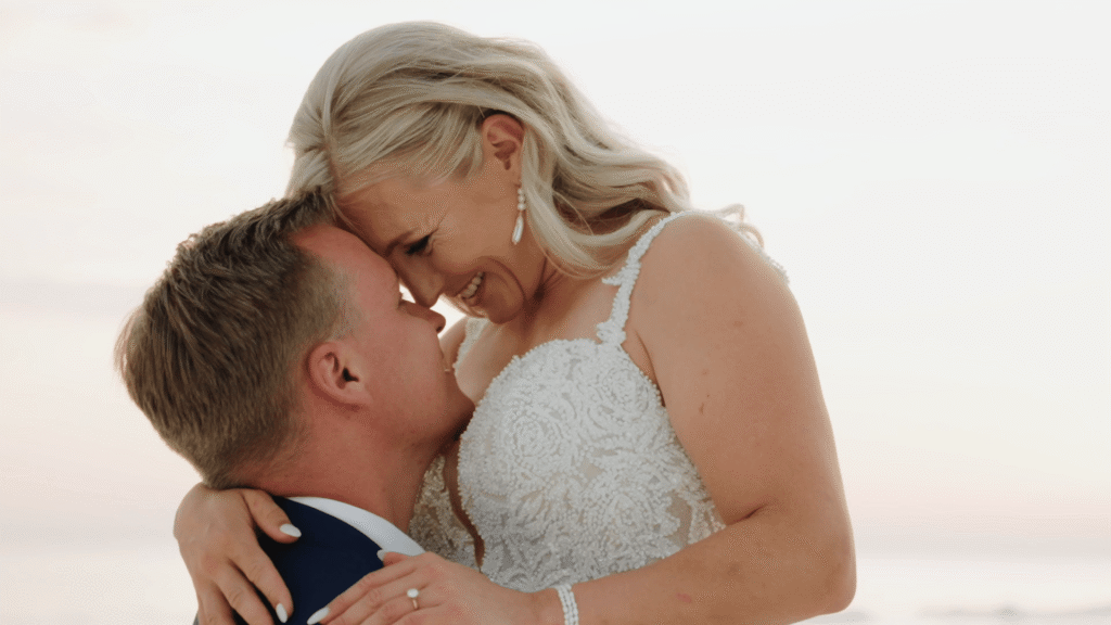 Bride and groom share a romantic embrace at sunset on Florida’s Gulf Coast, with the bride smiling joyfully in her lace gown.