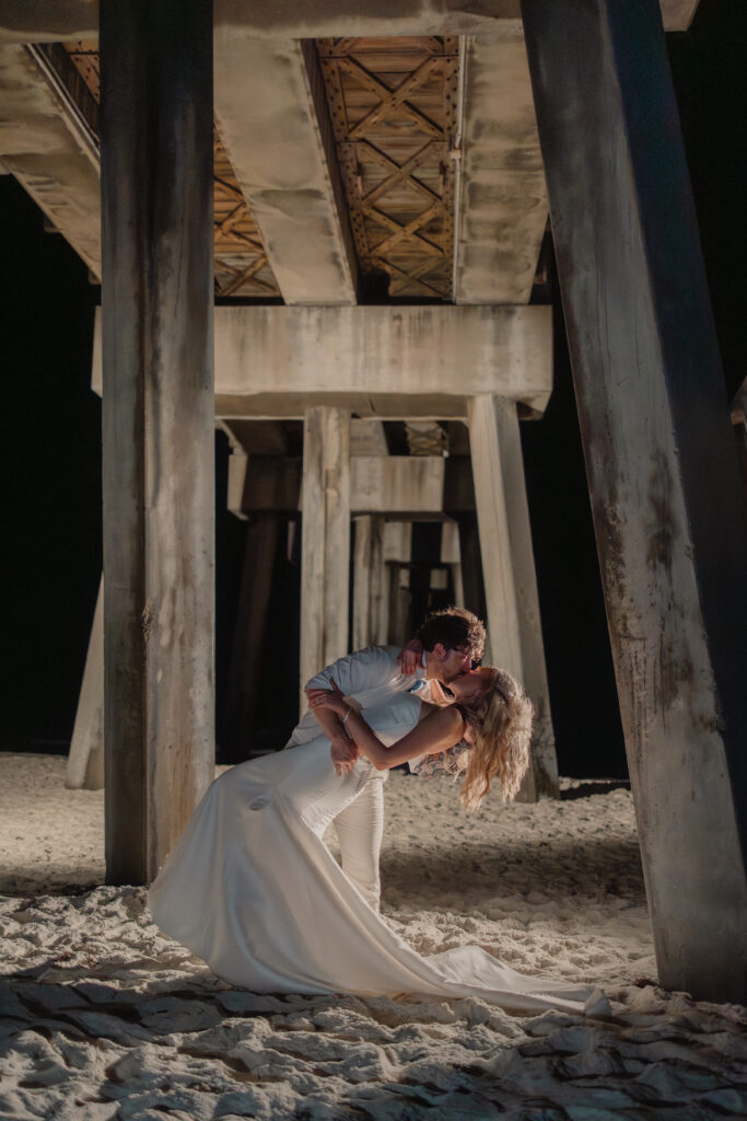 Bride and groom share a romantic kiss under a pier at night in Destin, Florida, captured by White Sands Weddings — highlighting the artistry and value of professional wedding photography.