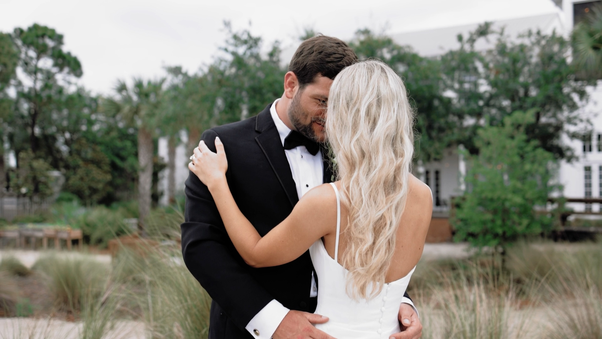Bride and groom share an intimate moment during their first look, with the groom in a black tuxedo and the bride in a white dress surrounded by greenery and soft natural light.
