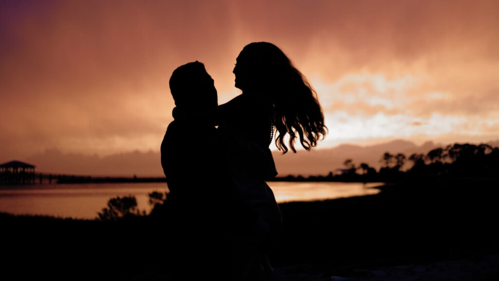 Silhouette of a bride and groom embracing at sunset by the water, with dramatic orange and purple skies in the background.