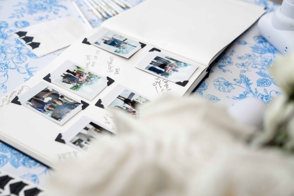 Close-up of a wedding guest book with Polaroid photos and handwritten messages, displayed on a floral blue and white tablecloth.