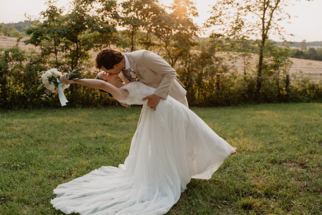 Bride and groom share a romantic sunset kiss, captured by White Sands Weddings with the bride’s lace gown flowing across the grass.