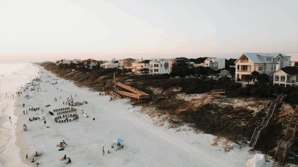 Aerial view of a beach wedding ceremony on 30A, with guests seated in a circle on the sugar-white sand and pastel homes along the coastline.