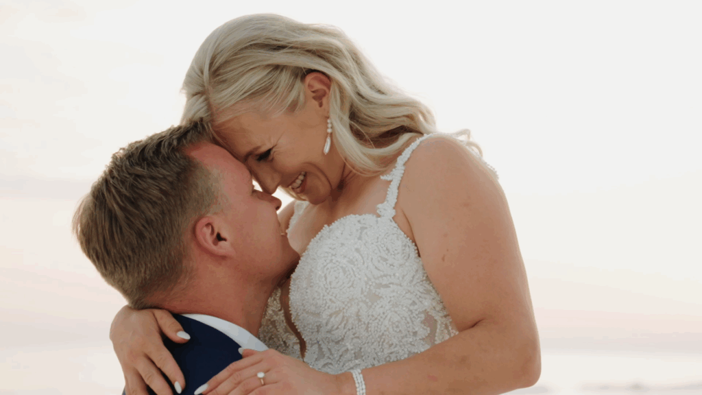 Bride and groom share a romantic embrace at sunset on Florida’s Gulf Coast, with the bride smiling joyfully in her lace gown.