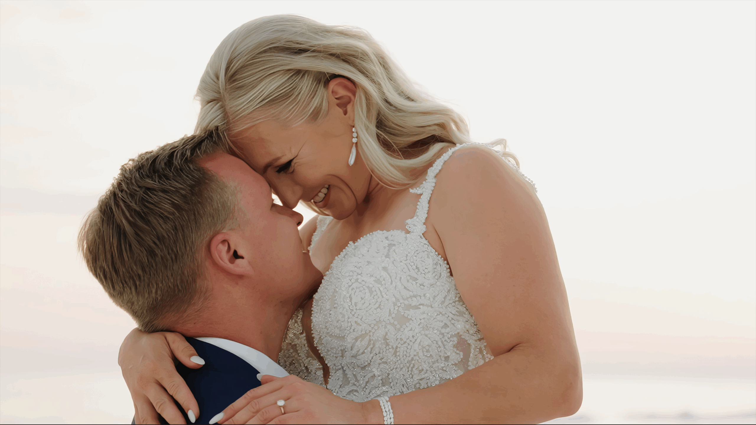 Groom lifts bride in a joyful embrace during golden hour beach wedding, with the couple smiling forehead-to-forehead under a soft pastel sky.
