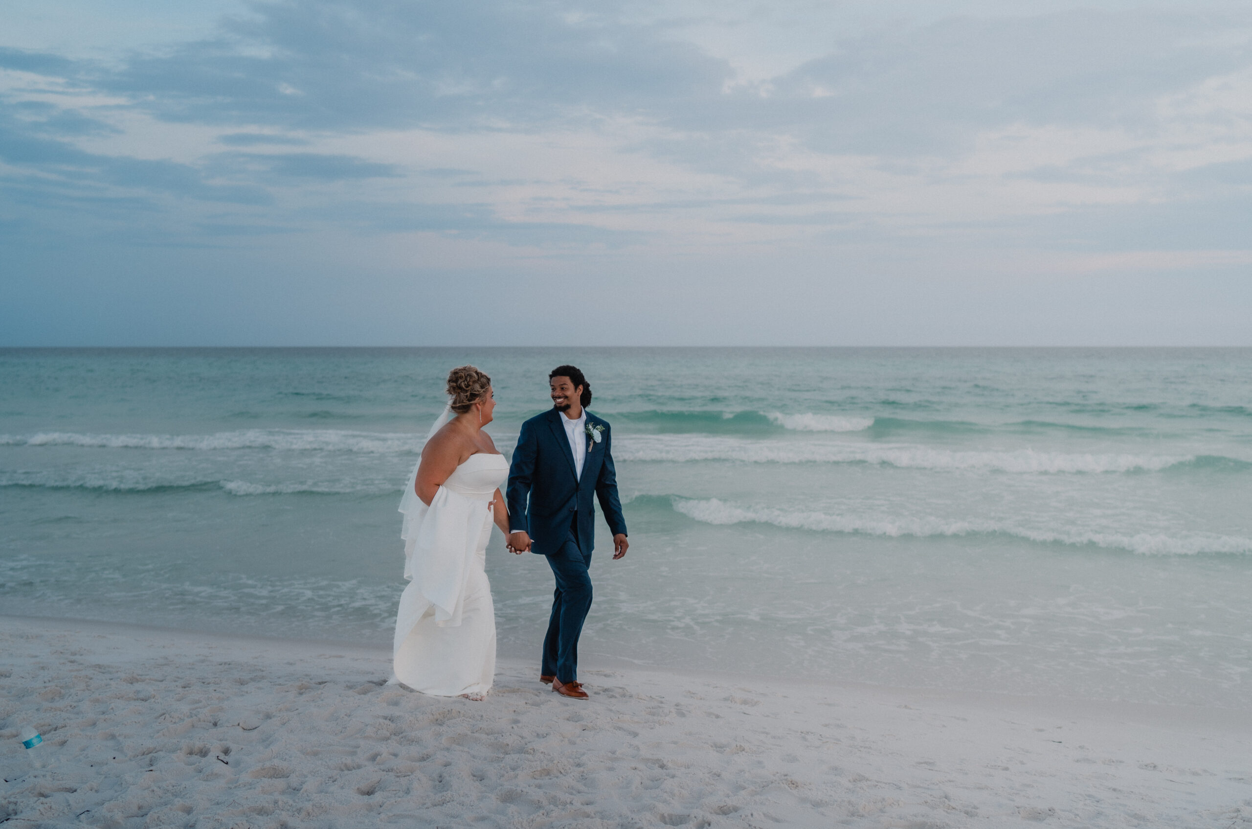 Bride and groom on a sugar-white sand Gulf Coast beach with turquoise Emerald Coast waters behind them, the groom in a navy suit.