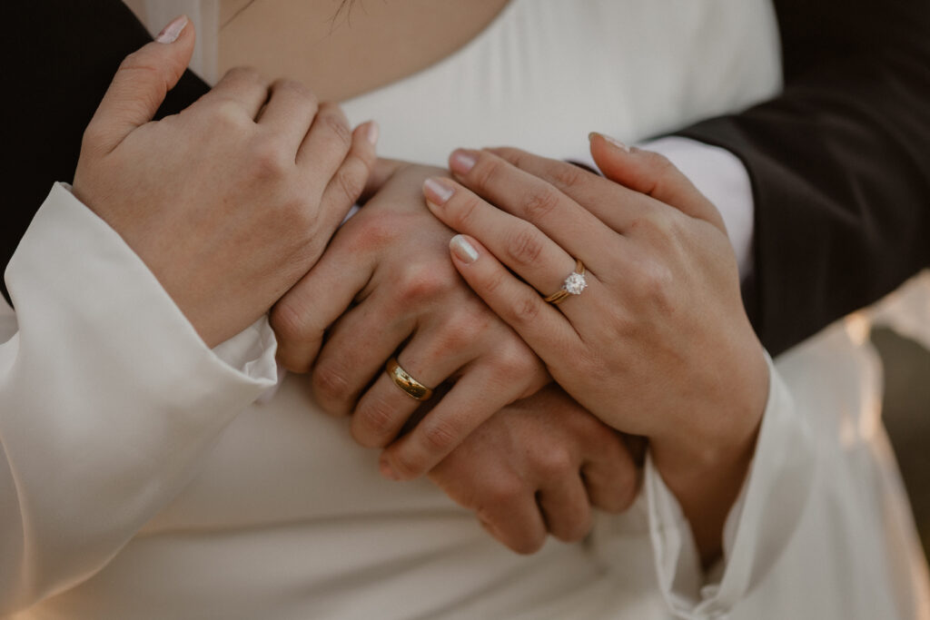 Close-up detail of bride and groom's hands with their wedding rings visible, the bride's lace cuff softly blurred in the foreground.
