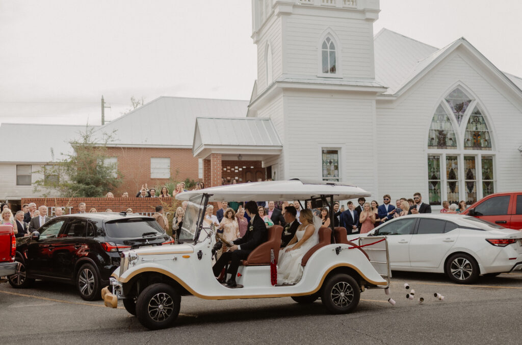 Bride and groom exiting their wedding ceremony in a vintage white car outside a historic Florida church with stained glass windows and a white bell tower.