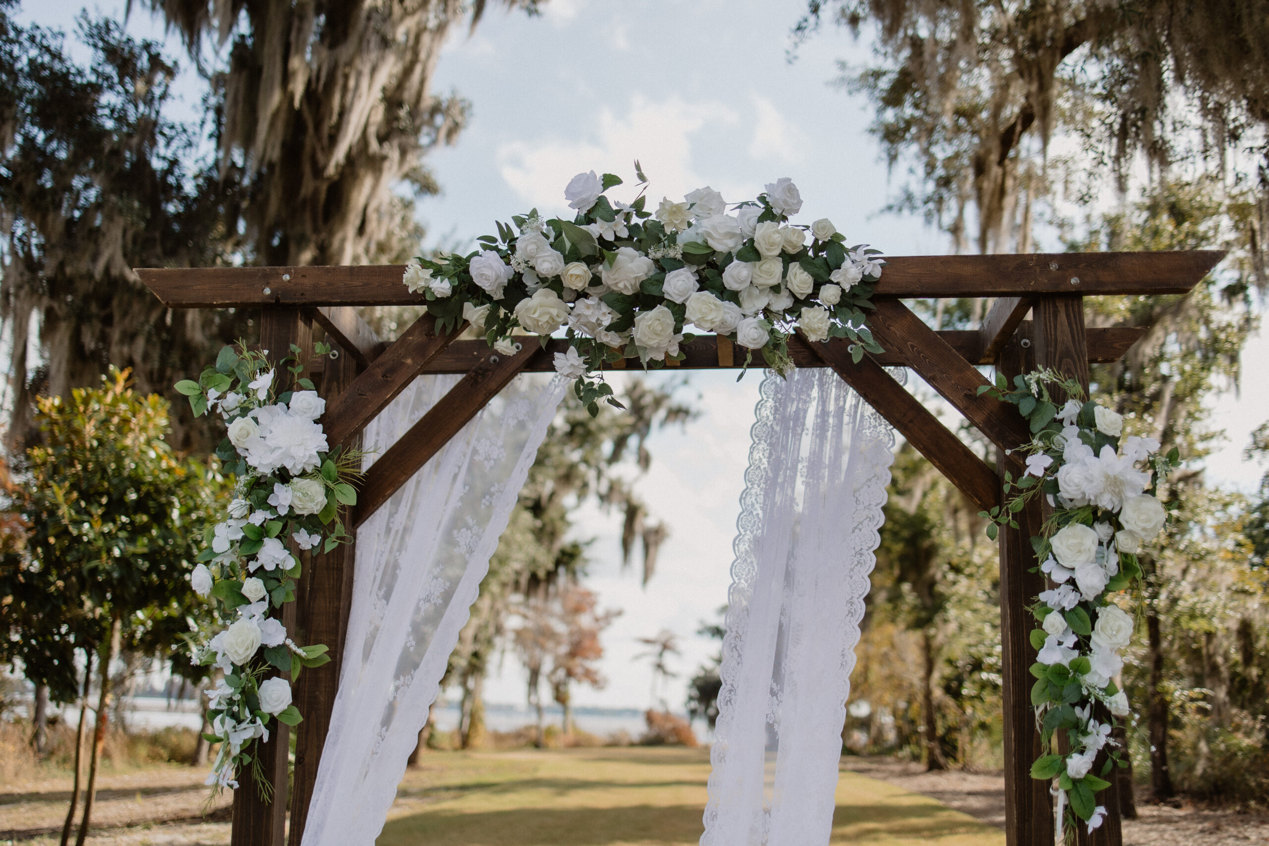 Rustic wooden wedding arch draped in flowing fabric and white florals on a sunlit Gulf Coast lawn ceremony setup.