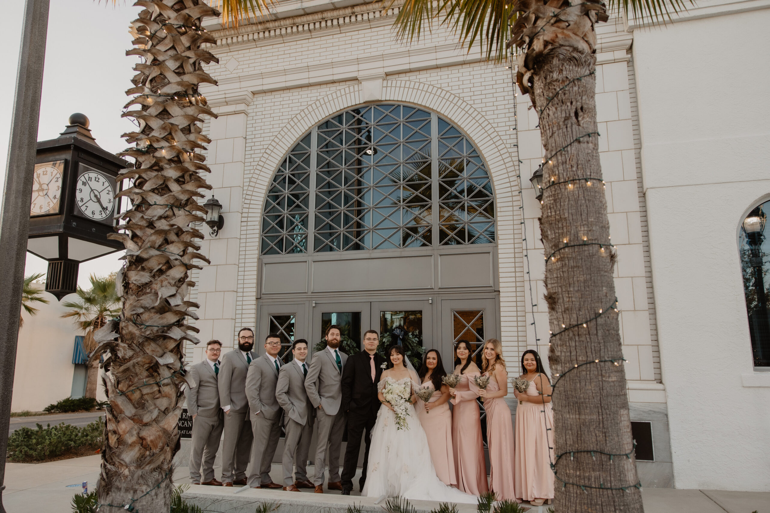 Gulf Coast wedding party of bridesmaids in blush gowns and groomsmen in grey suits posed in front of a historic arched-window building framed by tall palm trees at golden hour.