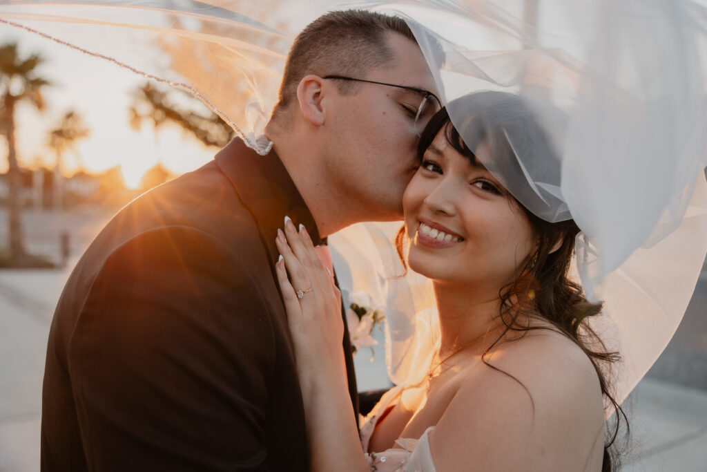 Groom kissing his bride's cheek during a golden-hour portrait session on the Emerald Coast, with warm sunset light wrapping around the couple.